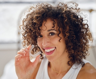 Woman with curly brown hair smiling while holding dental floss in bright dental office setting.