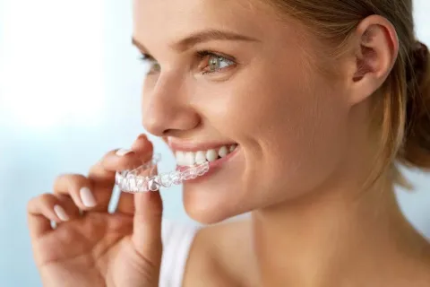 Woman holding clear aligner near her smile against white background, demonstrating orthodontic treatment.